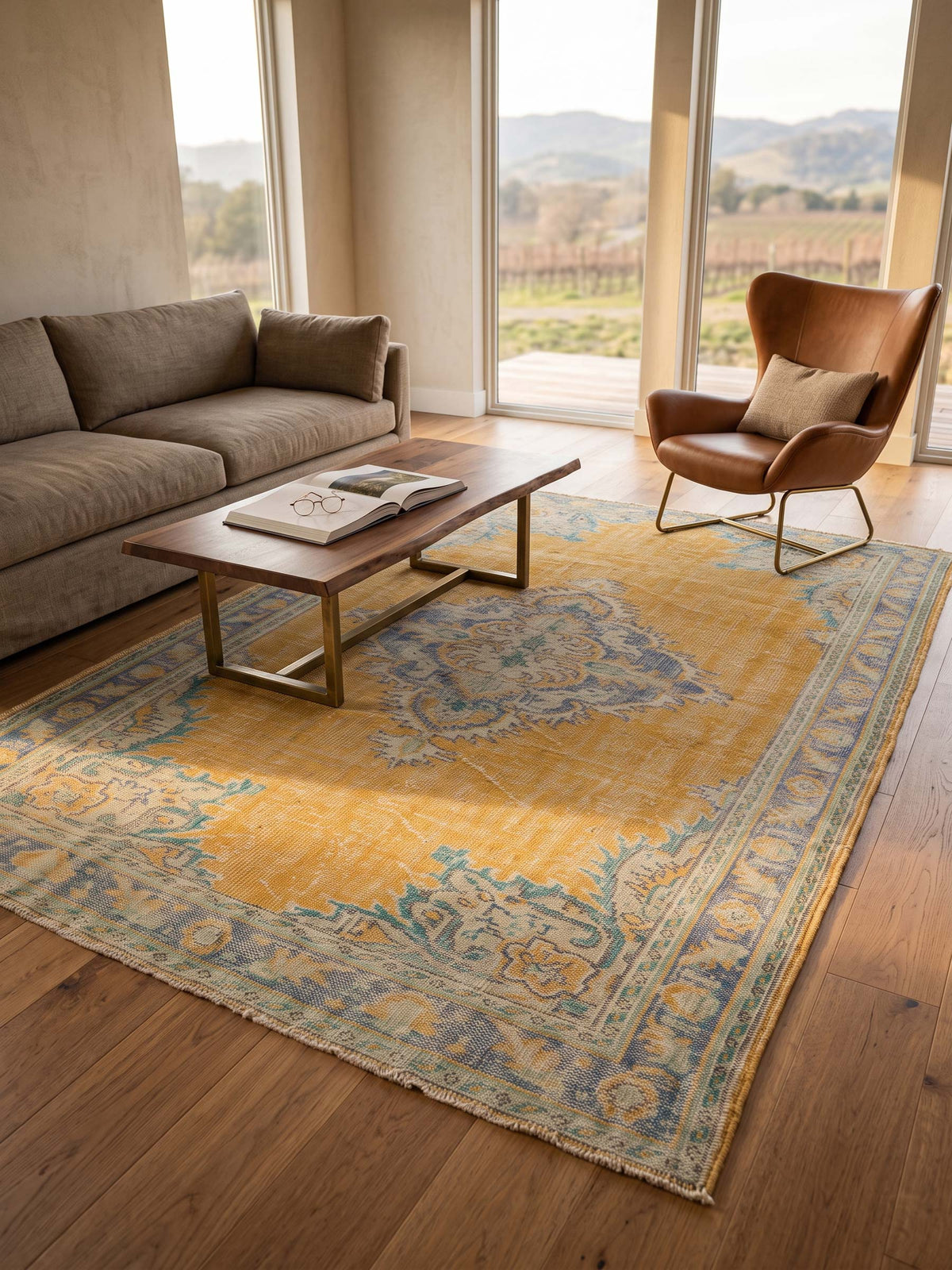A large traditional-style area rug in mustard yellow, blue, and beige tones, styled in a modern living room with a brown leather sofa, a wooden coffee table, and a brown armchair near large windows overlooking a vineyard.